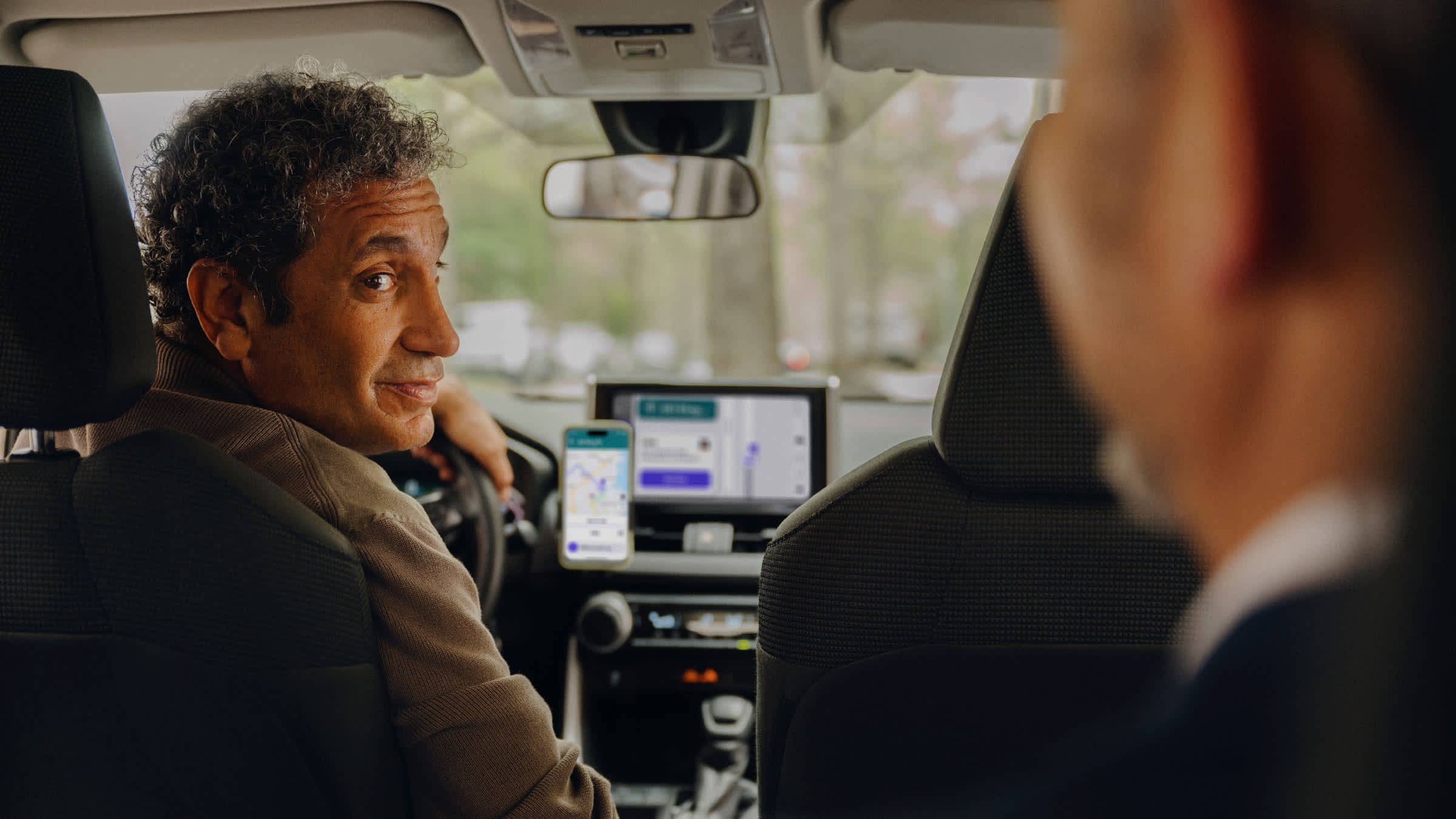 Driver in brown jacket looking at passenger, with Lyft app and navigation screen visible in car.