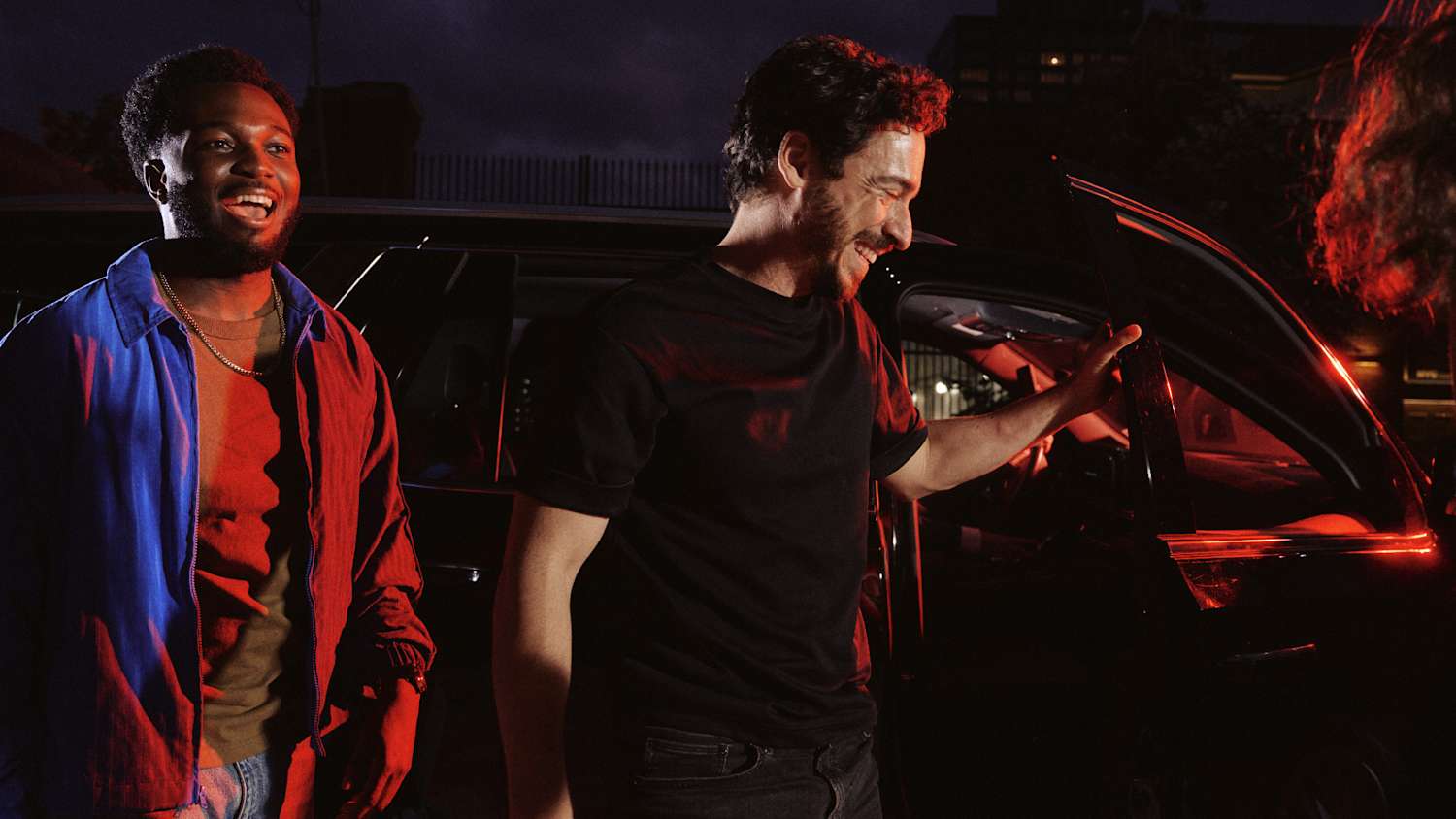 Two men smiling and standing by an open car door at night, illuminated by red and blue lights.