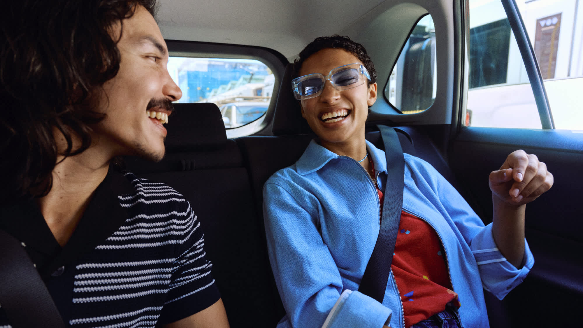 Two people smiling and talking in the backseat of a car, both wearing seatbelts and casual clothes.