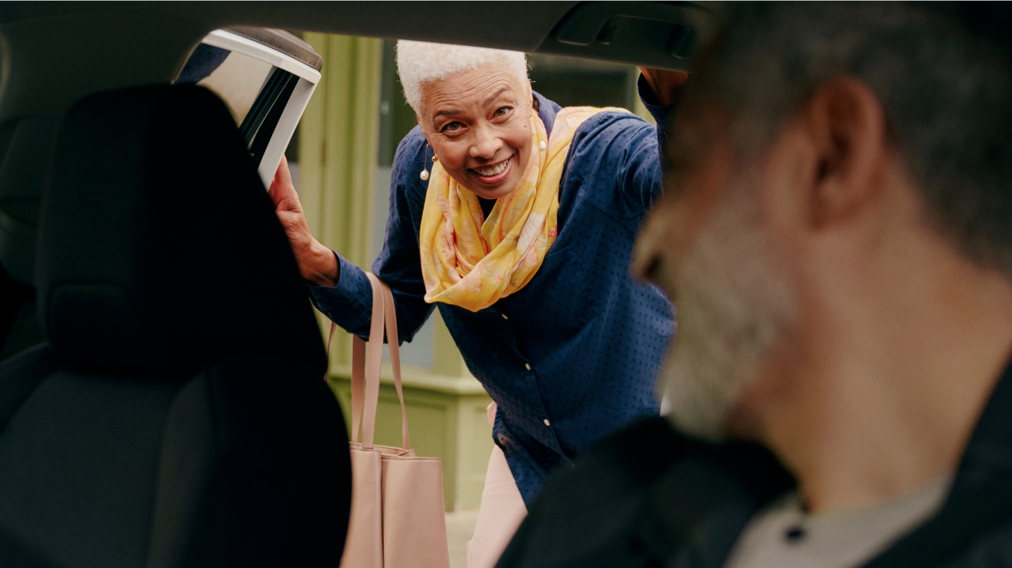 Smiling older woman with a yellow scarf leans into a car, holding a tote bag, while a man looks back at her.