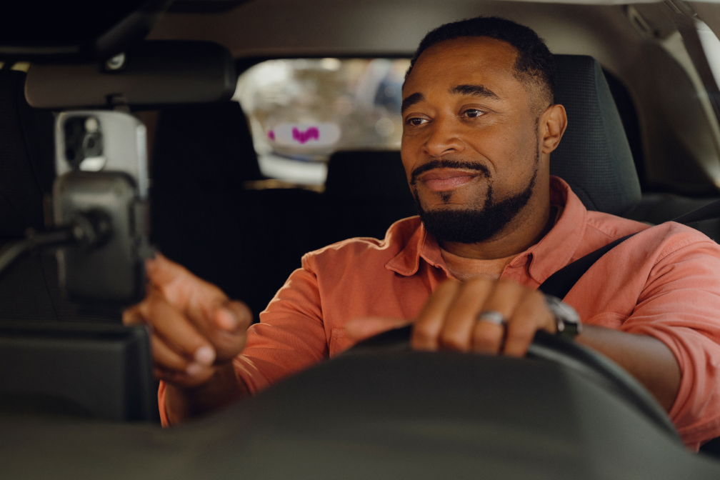 Smiling man in a coral shirt drives a car, touching a phone mounted on the dashboard, with a Lyft sticker visible.