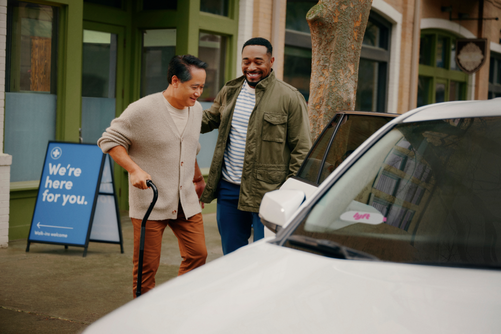 Two men smile as one helps the other, who uses a cane, get into a white car on a city street near a blue sign.