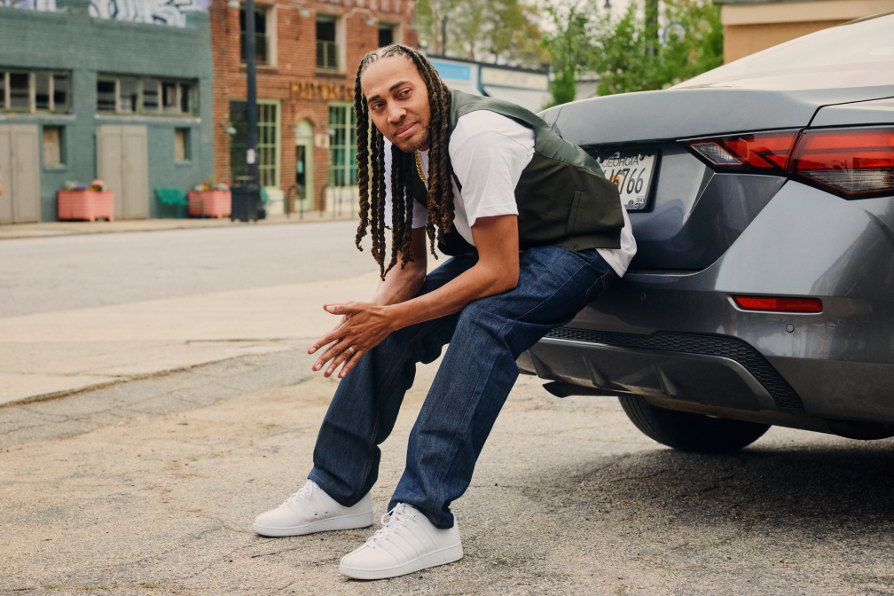Man with long braids, white sneakers, and casual clothes sitting on the back of a gray car on a city street.